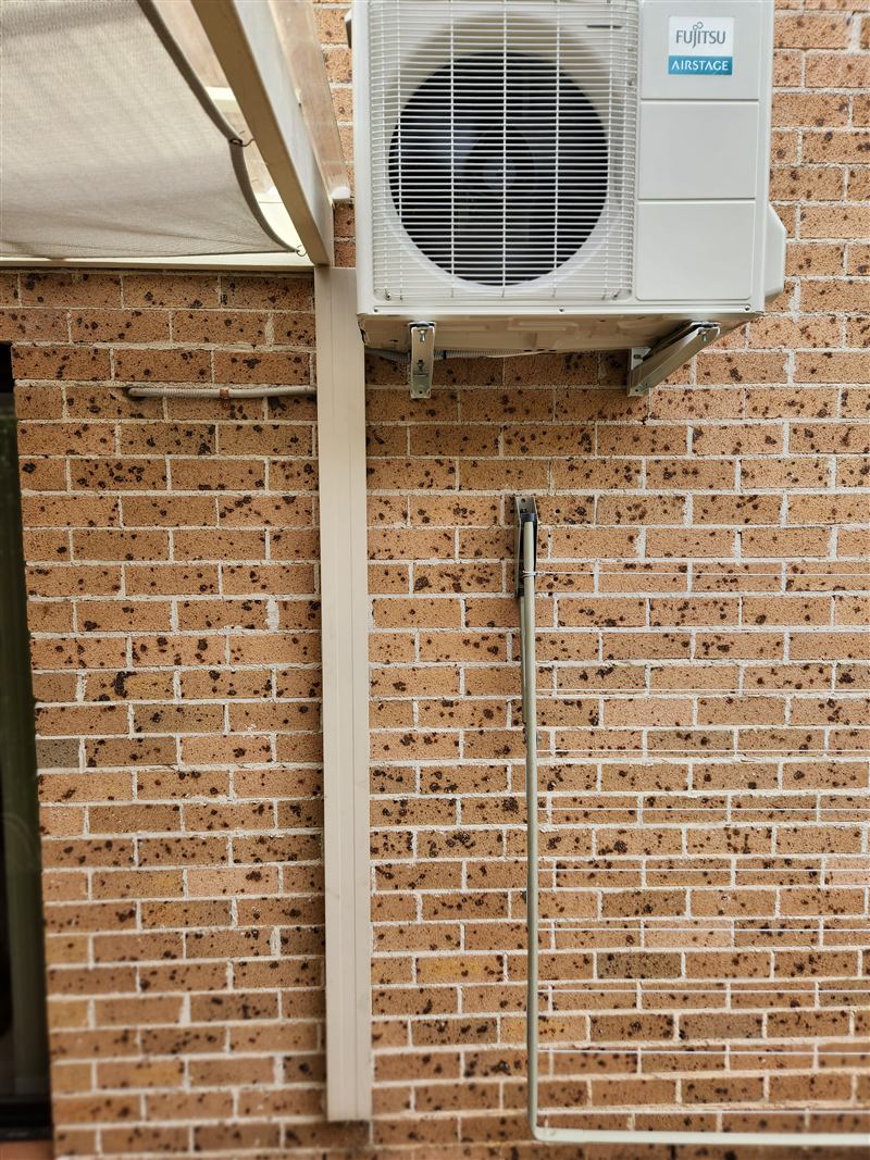 A reverse cycle outdoor unit mounted on wall brackets at a brick home, with neat pipe cover running down the wall